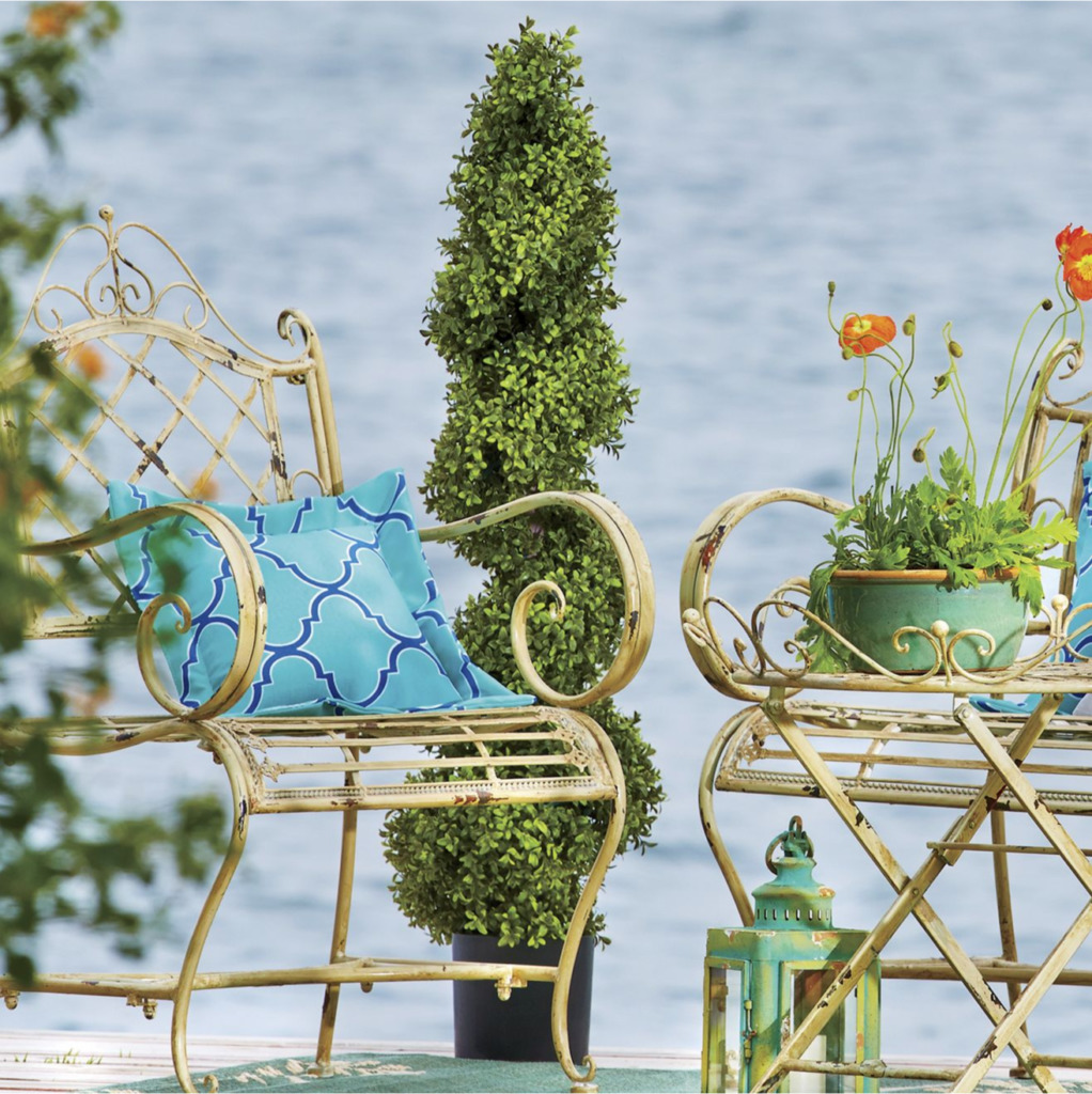A spiral boxwood topiary poolside, beside two Victorian metal chairs, a blue pillow, and potted orange flowers.