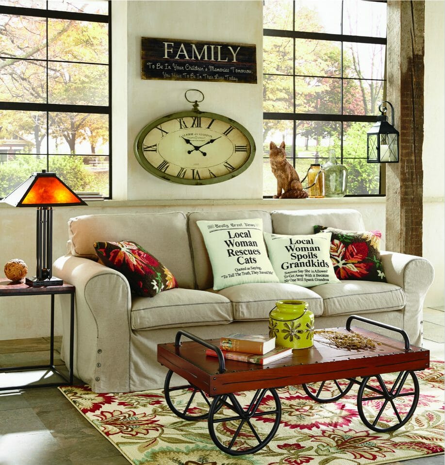 Ivory sofa with floral and versed pillows, a wood and spoke wheels cart table, a floral area rug, and an oval wall clock.