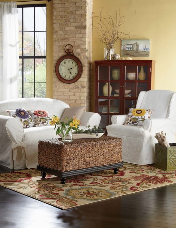 White slipcovered chair and sofa, seagrass storage coffee table, glass doors cupboard, sunflower pillows, and a wall clock.