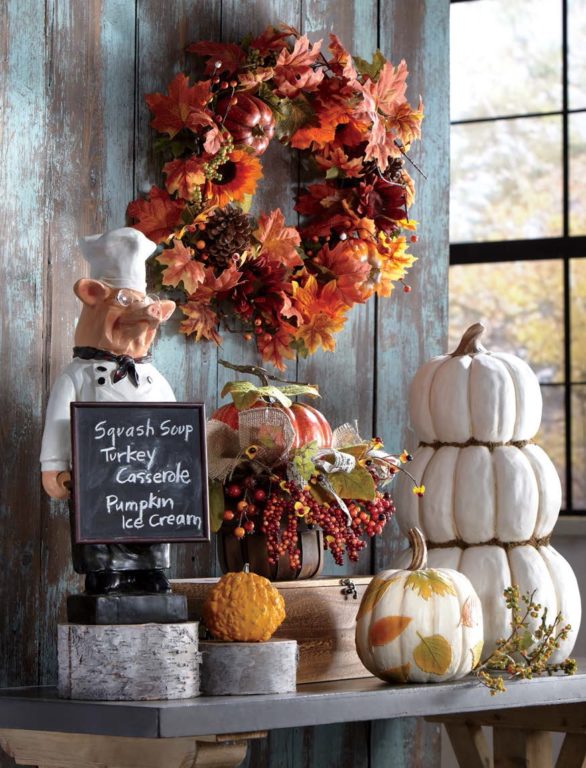 A Fall wreath above a pig chef holding a chalkboard, and white and orange pumpkins on a rustic table.