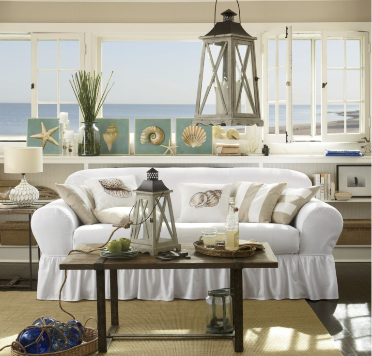 Nautical themed living room with a white sofa, shell pillows and plaques, lanterns, blue glass floats, and an ocean view.