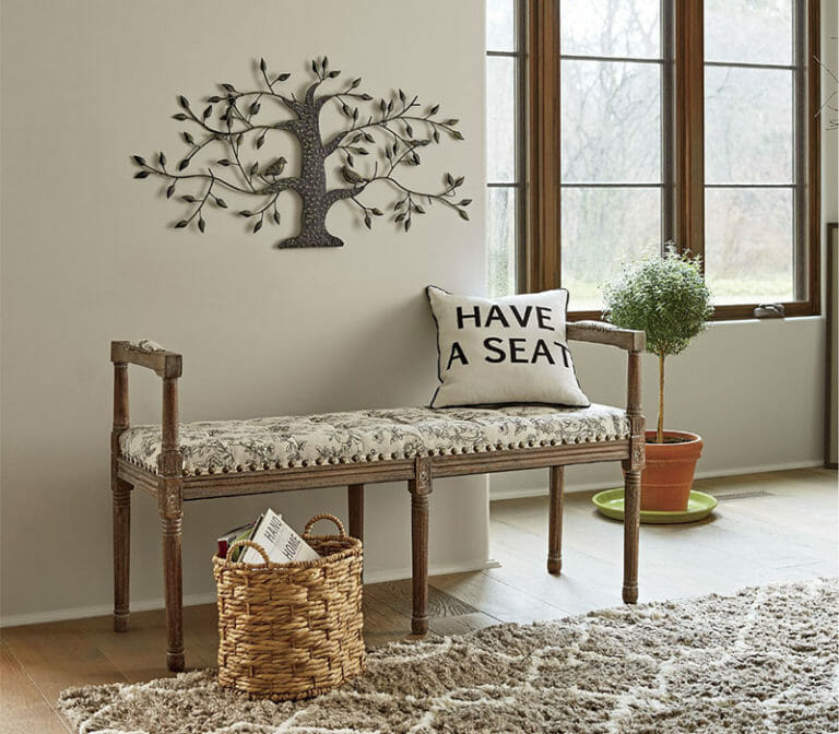 An entryway with a metal tree of life above a floral bench, a magazine basket and a potted topiary on the floor.