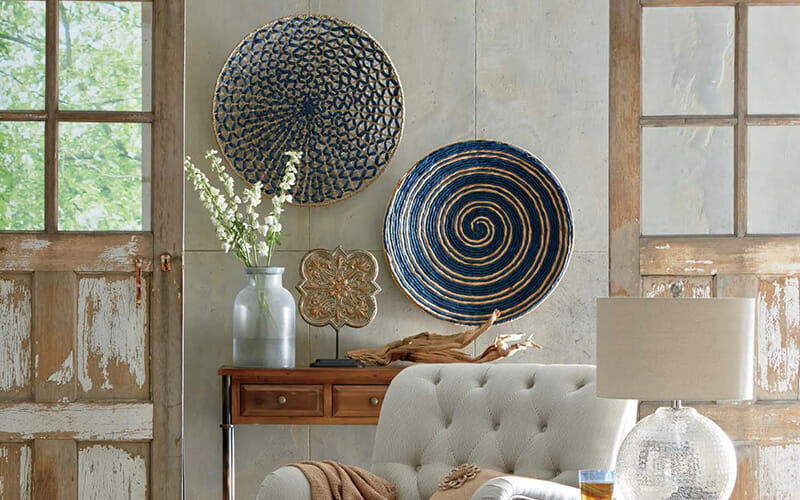 Two large flat blue and tan baskets hung on a wall above a console, with a white tufted armchair, and glass table lamp.