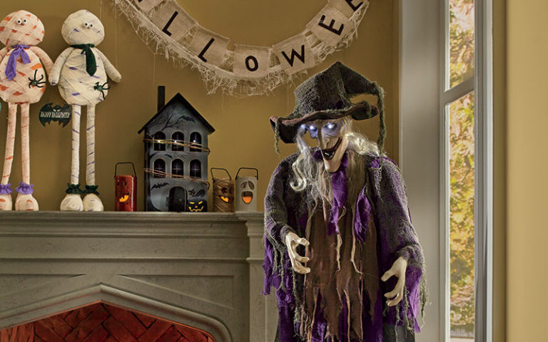 Skeleton witch with glowing eyes dressed in rags in front of fireplace and ghost candles with other Halloween décor