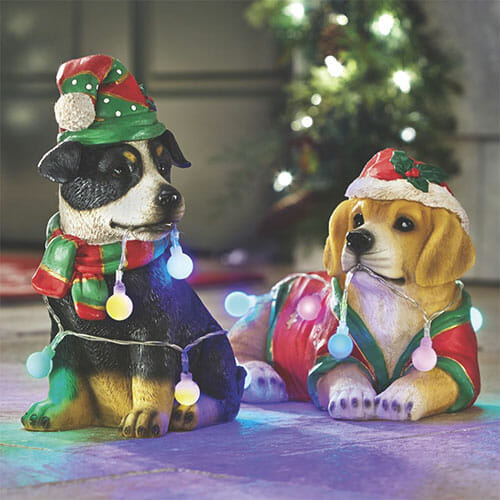 Two dog statues in Christmas hats and scarves, with lit outdoor string lights wrapped around them.