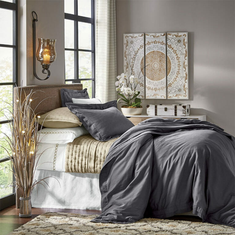 A bed with layered gray, tan, and white bedding, a lit wall sconce, tryptic baroque wall panels, and a potted white orchid.