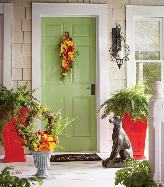 A green front door with a yellow and orange cosmos wreath, two tall red planters with ferns, and a black dog statue.