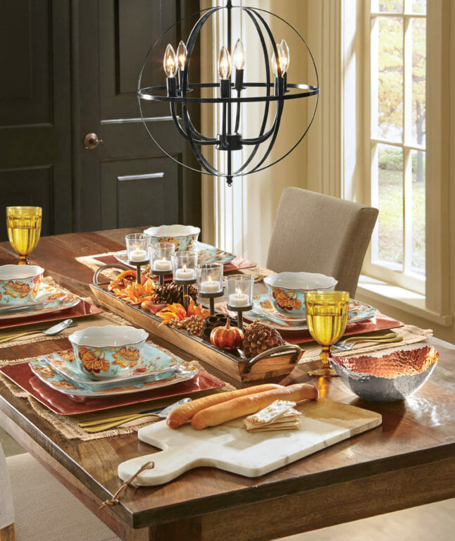 A table with a lit candle Fall centerpiece, teal and orange floral dinnerware, amber goblets, and crusty bread on a board.