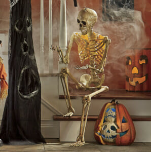 A tall posable skeleton sitting on a staircase, with a black haunted tree stump, lit pumpkins, and swirling smoke.