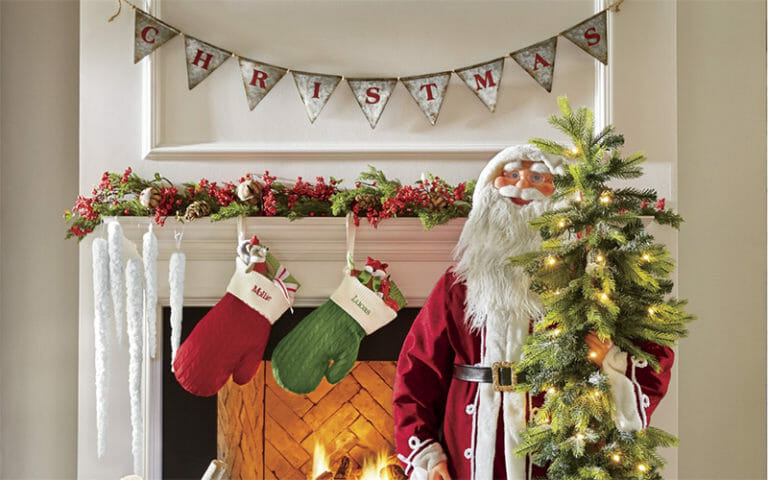 Green and red display of wrapped presents, a tall Santa with a tree by a lit fireplace hung with mittens.