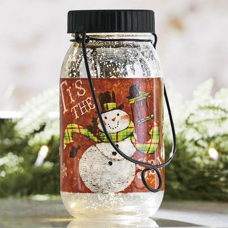 A lit glass jar of a painted happy snowman holding a lantern, with a black lid and hanger.