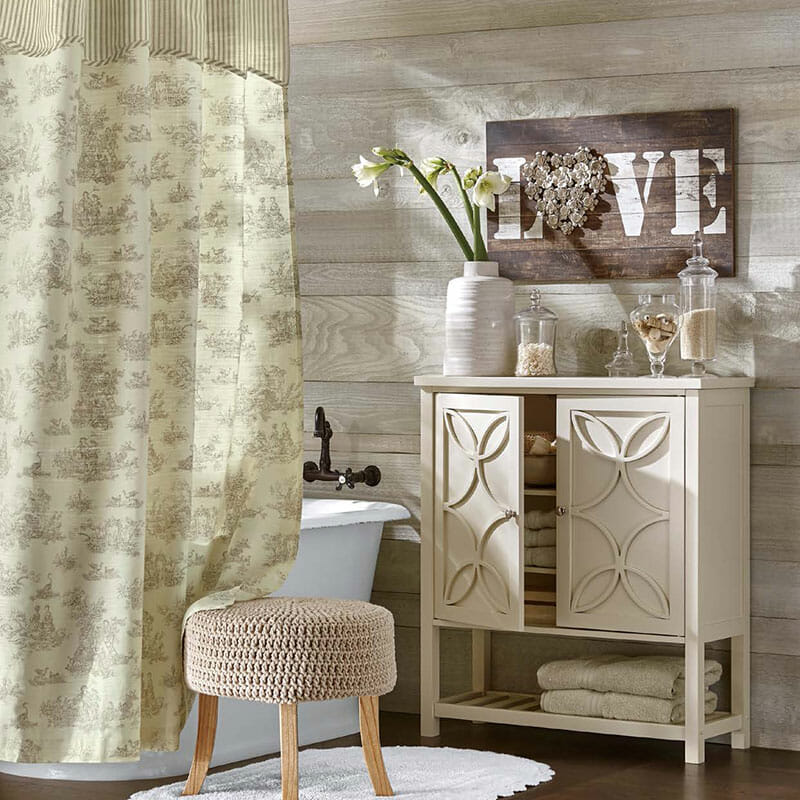 A beige and gray toile shower curtain around a freestanding tub, an ivory cabinet with towels, and a LOVE wood plaque.