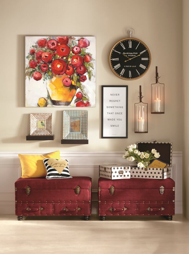A large red floral canvas, a round wall clock, two lit sconces, and a framed verse above two burgundy traveler's trunks.