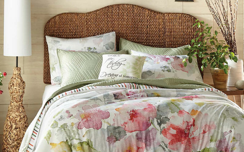 A watercolor floral comforter set with a seagrass headboard, a floor lamp with a woven base, and a potted green plant. 
