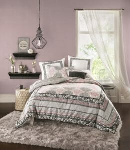 Mauve, black and white comforter set with a beige glazed side table, gray shag area rug, and a sheer gray curtain.