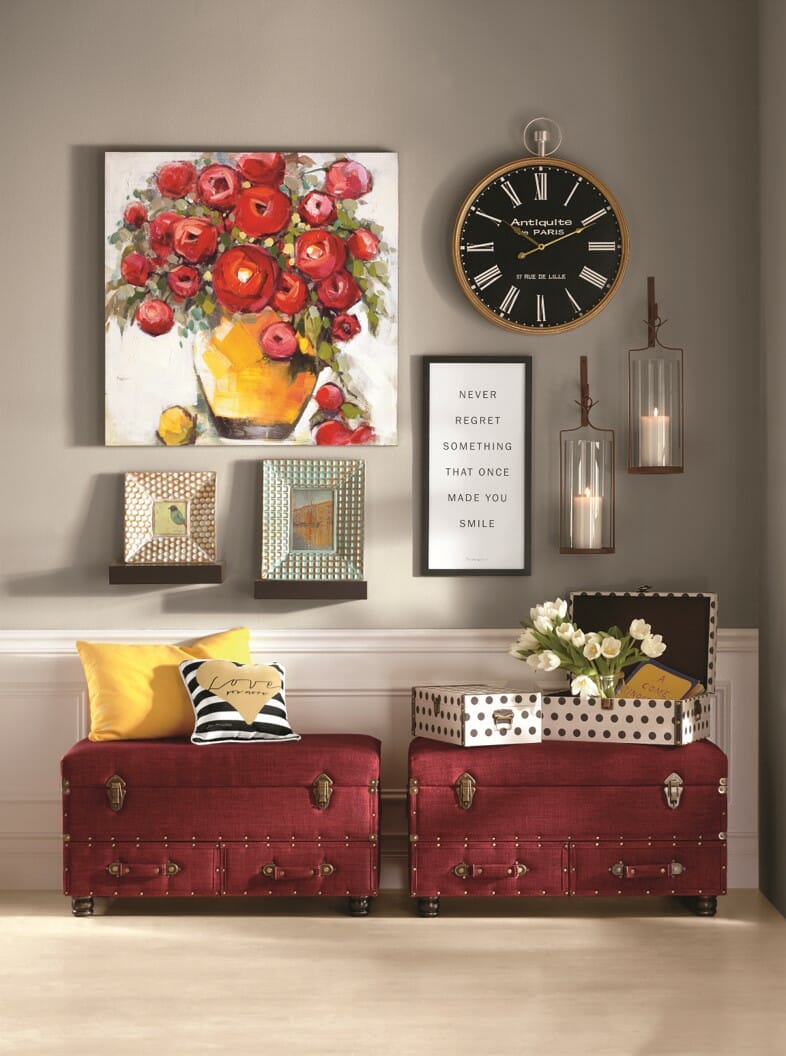 A large red floral canvas, a round wall clock, two lit sconces, and a framed verse above two burgundy traveler's trunks.