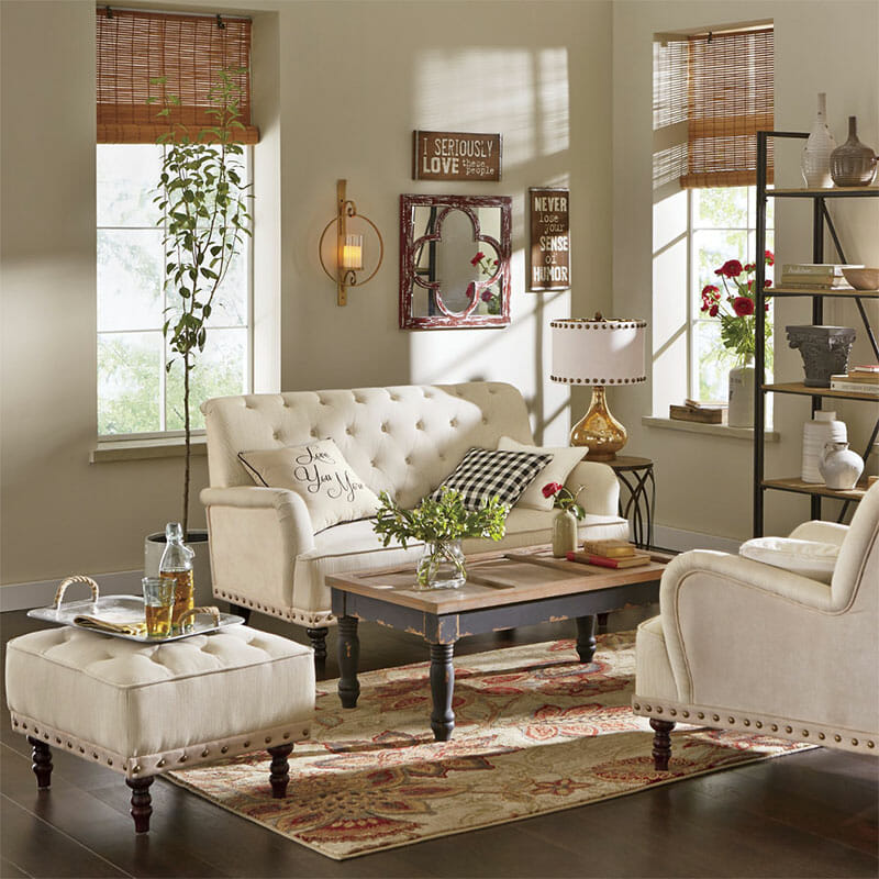 Living room with ivory tufted sofa, chair and ottoman, a floral rug, a framed mirror, a lit sconce, and a beverage tray.