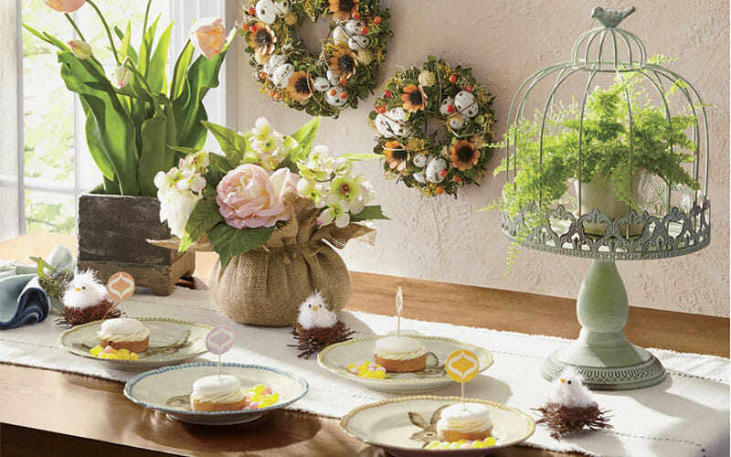 A table set for Easter with fluffy white chicks on nests, a pedestal bird cage with a fern, potted tulips and frosted cupcakes.