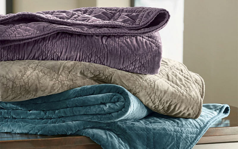 A stack of three folded solid color quilts, in aubergine, tan, and teal.