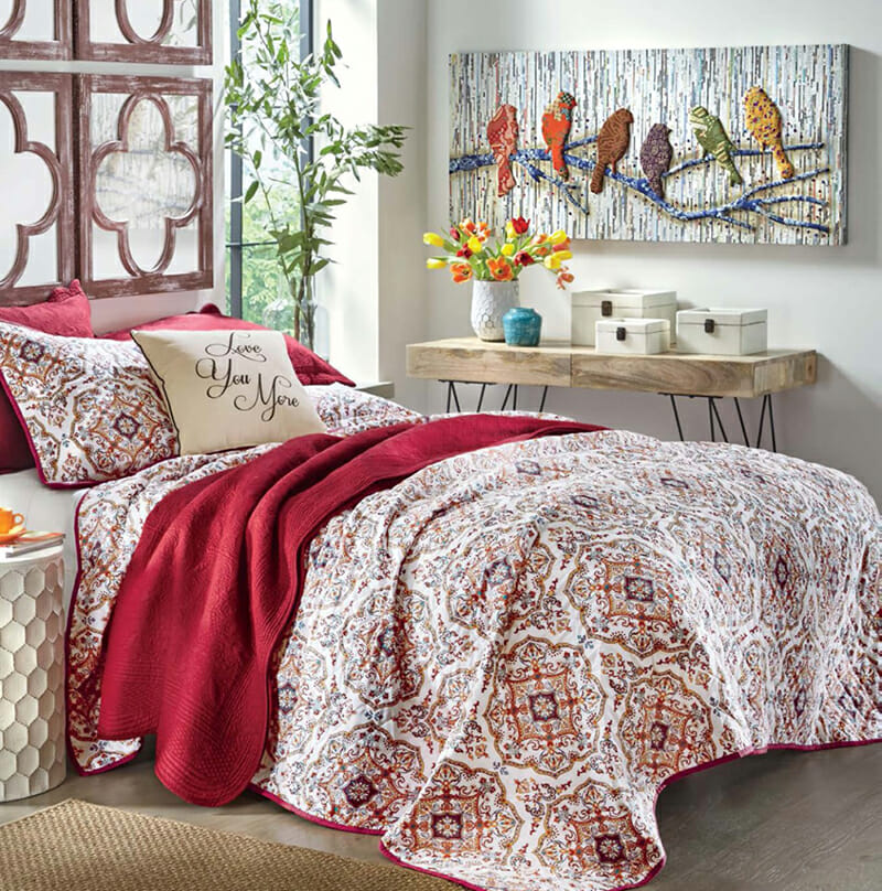 A solid red quilt set layered with a baroque red and white quilt set, and a 3D wall canvas of multicolored birds.