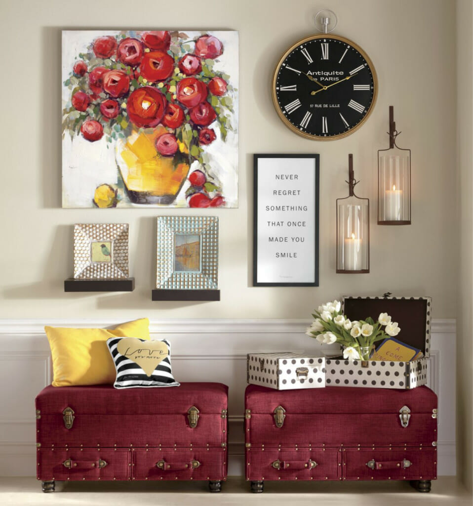 Two red trunk benches with pillows, a large canvas of red roses in a yellow pot, a round black clock, and two lit sconces.