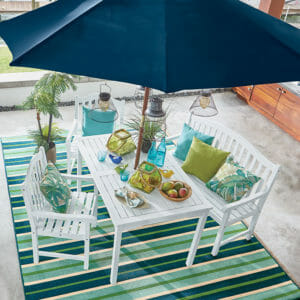 A white patio set with shades of aquamarine in an umbrella, striped rug, pillows, and bottles on the table.