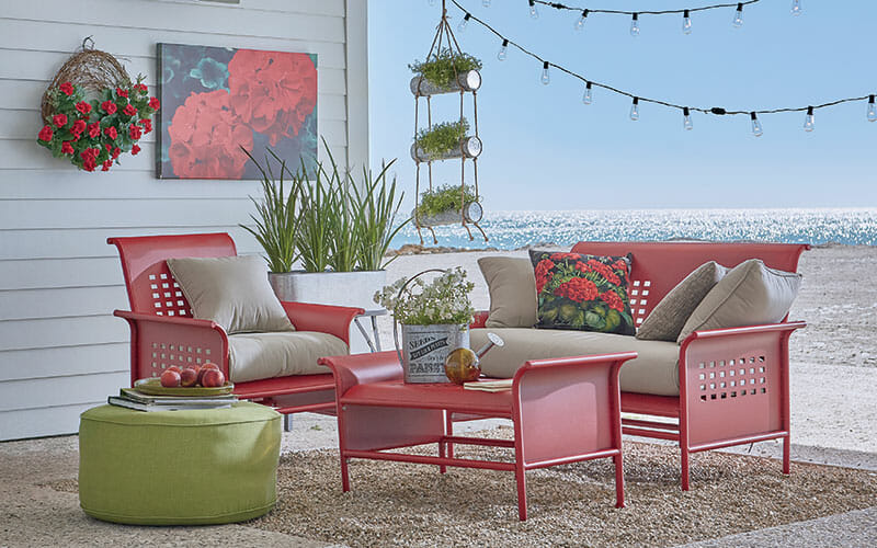 Retro red metal patio set with beige cushions, a large red geranium canvas and wreath, green plants, and an ocean view.