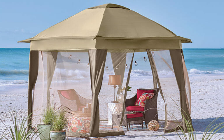 A tan gazebo with screens on a beach, with patio chairs with red cushions, a table lamp, and a string of lights.