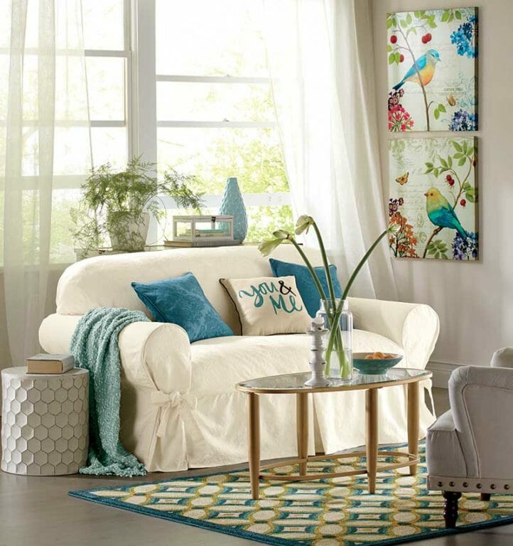 A white slipcovered sofa with teal pillows and throw, with two multicolor bird canvases on the white walls.