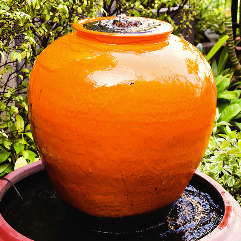An orange bubbling jar fountain by green bushes in a garden.