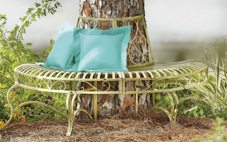 A vintage half circle beige metal bench with a scroll top placed around a tree trunk, holding two teal pillows.