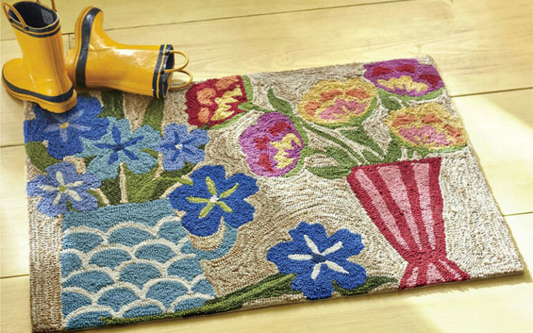 A colorful floral hooked rug in blues, pinks, and yellow on a wood floor, with a pair of yellow rain boots.