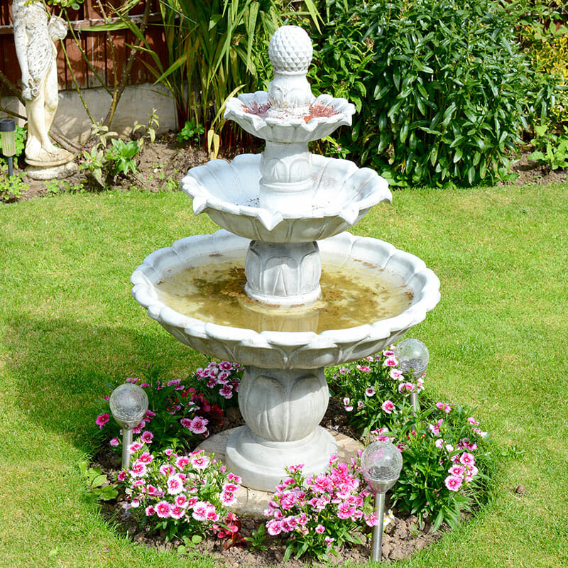 A three-tier cement water fountain in a garden, surrounded by pink and white dianthus and solar globes.