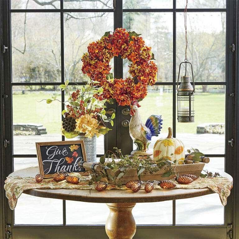 A round table by a window, covered with a Fall floral table runner, wood tray with a pumpkin, a rooster, and vased flowers.