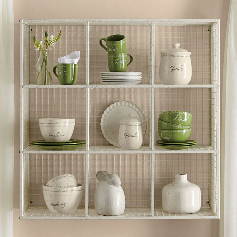 A white Farmhouse divided wire shelving unit, holding green and ivory dinnerware, a ceramic rabbit, and vased flowers.