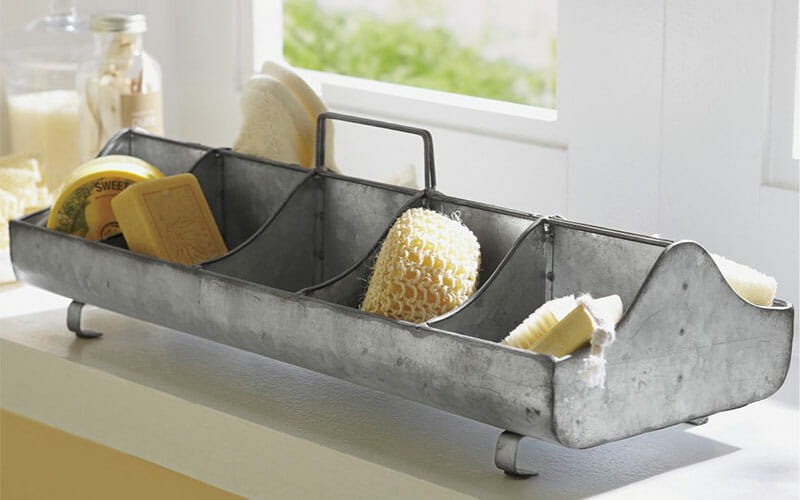 A rustic galvanized metal tray on a countertop, with soaps, a sponge and brushes in the divided sections.