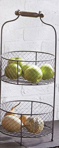 Two stacked round wire baskets with a wood handle, with green apples on top and gourds on the bottom.