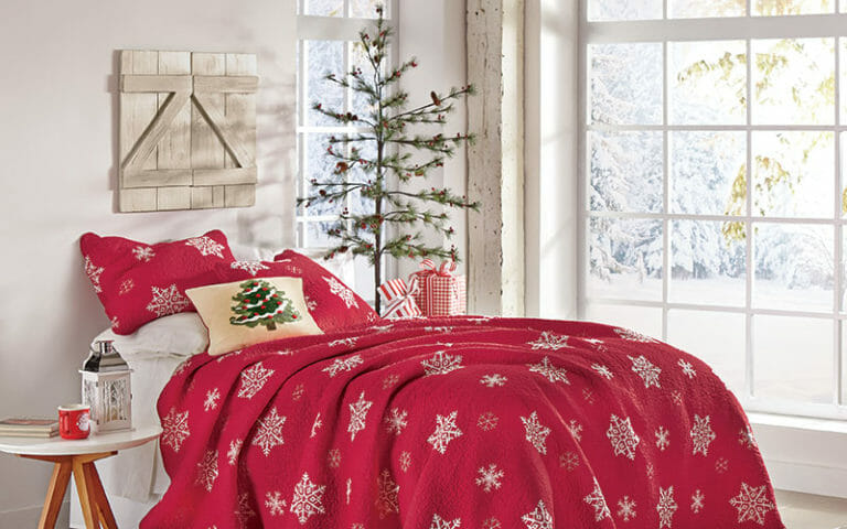 A red quilt and shams with white snowflakes, a small pine tree with presents, and a snowy view out the large paned windows.
