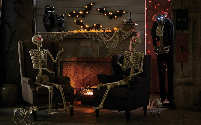 A dark room with a lit fireplace, two skeletons in black chairs, a dog and cat skeleton, and pumpkin lights on the mantle.