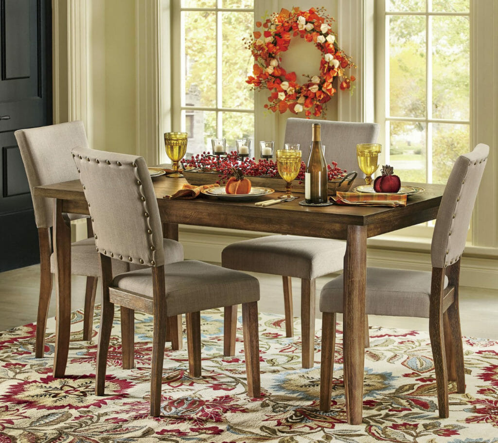 A wood table and four upholstered chairs, set for a Thanksgiving dinner, with a floral area rug and a Fall wreath.
