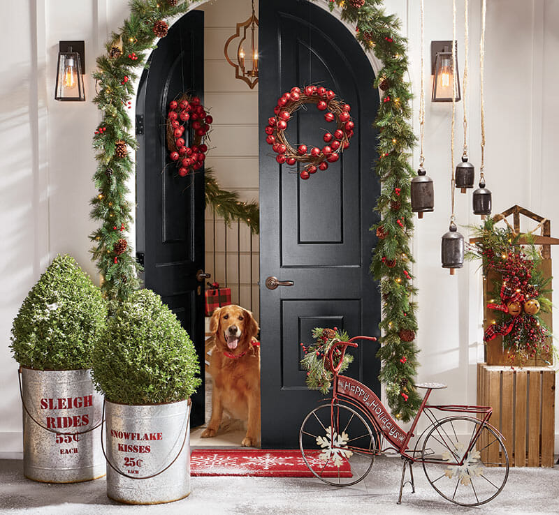 Two red jingle bell wreaths on open double doors hung with pine garland, with a dog looking out.