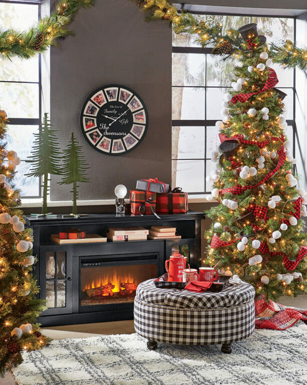 A lit electric fireplace by a Christmas tree decorated with black top hats, snowballs, and red check ribbon.