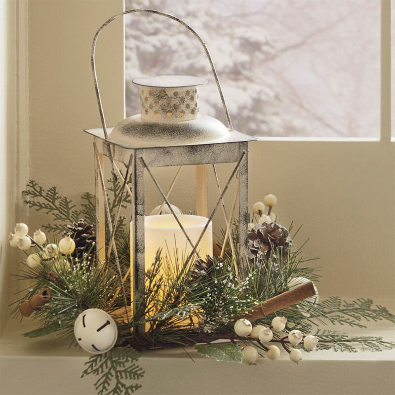 A metal lantern by a snowy window view, with a lit white candle, pine, white berries, pinecones, and cinnamon sticks.