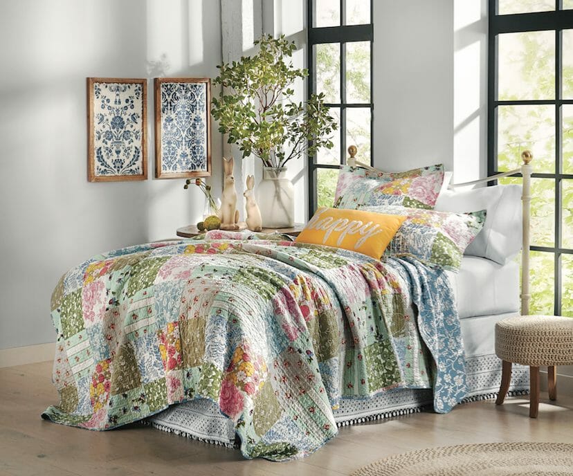 A multicolored floral squares quilt and shams, with a yellow happy pillow, two rabbit figurines, and a crochet stool.