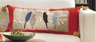 A long red pillow with gold tassels, with an applique of four blue bird silhouettes on pom pom wires.