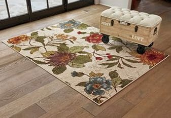 A beige area rug with large botanical flowers in burgundy, orange, blues, and greens, and a wood cart with a tufted seat.