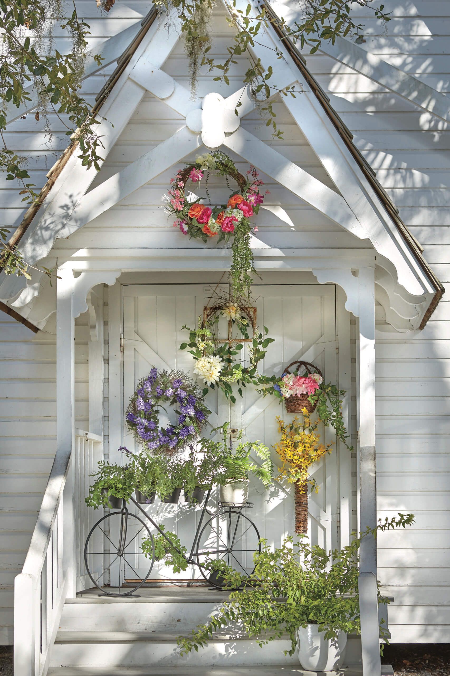 Five colorful Spring wreaths hanging on a white country schoolhouse entrance with an iron bicycle plant stand holding ferns.