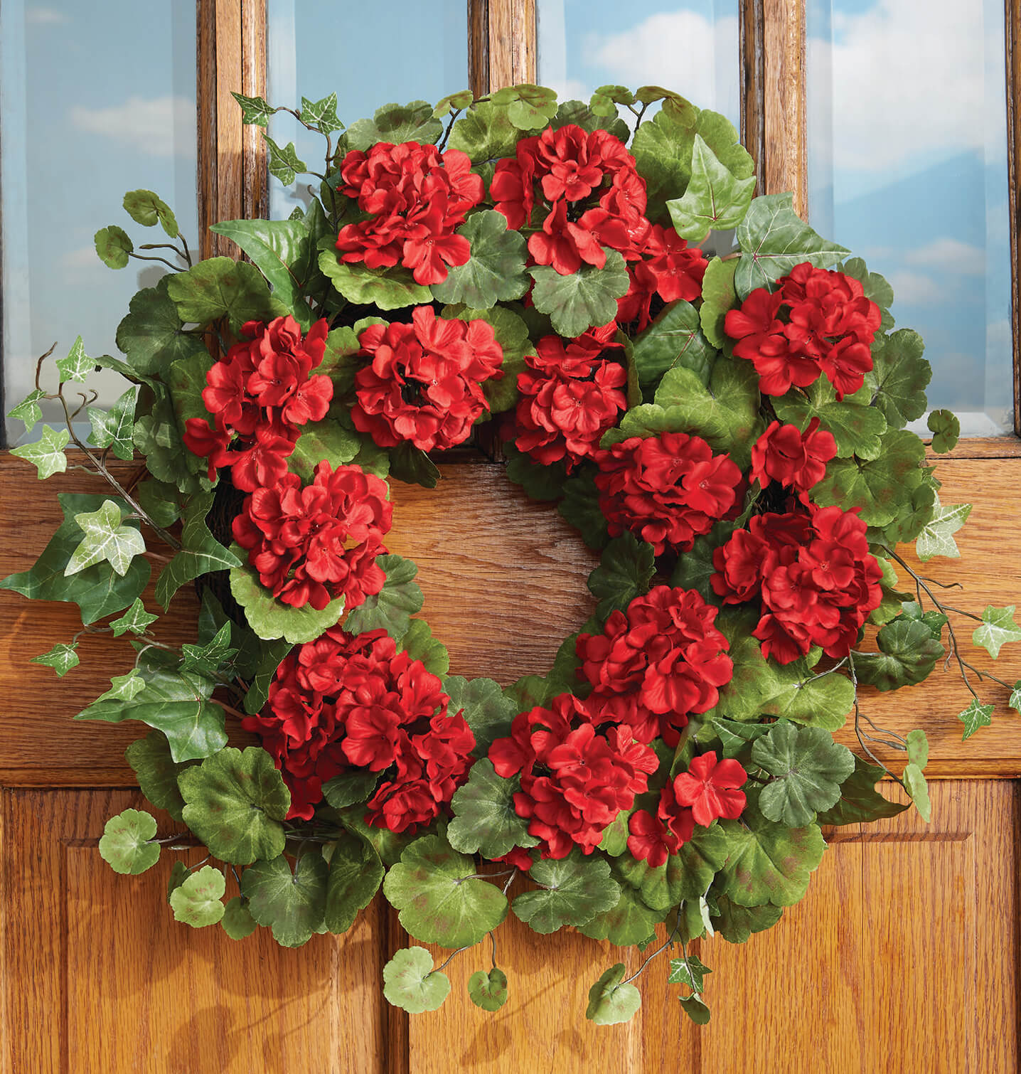 A round red geranium wreath with ivy hung on an oak door with paned windows.