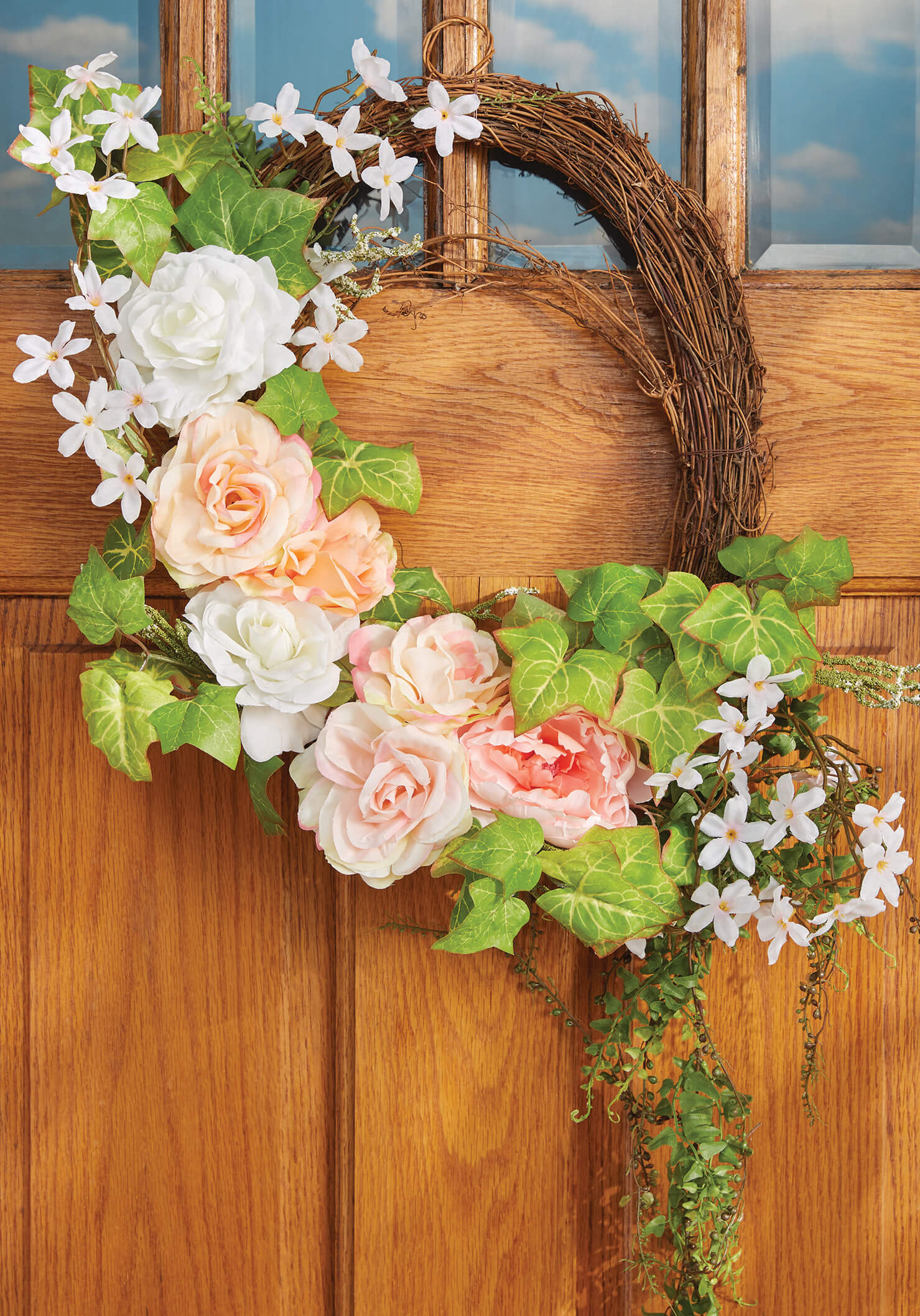 A round grapevine wreath of peach and white roses, ivy and white flowers hung on an oak door with paned windows.