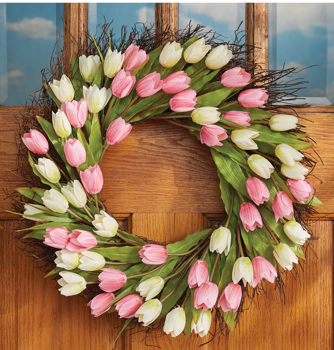 A round twig wreath of many pink and white tulips hung on an oak door with paned windows.